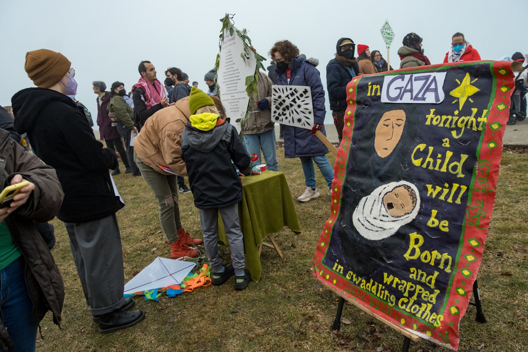 Protesters work at a craft table next to a sign that says "Tonight, in Gaza, a child will be born and wrapped in swaddling clothes."