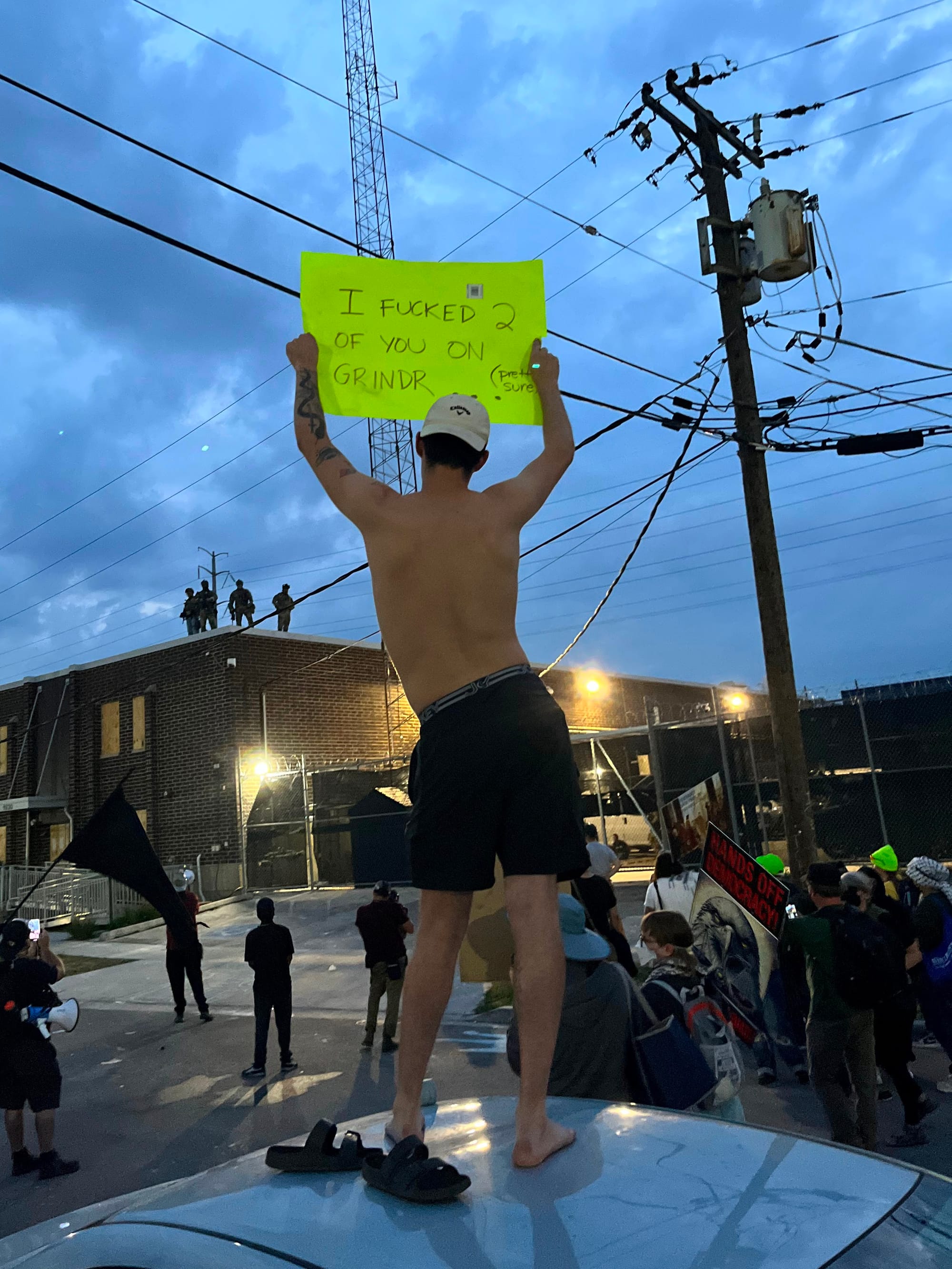 A man standing on the roof of a car holds a sign that reads: "I fucked two of you on GRINDR."