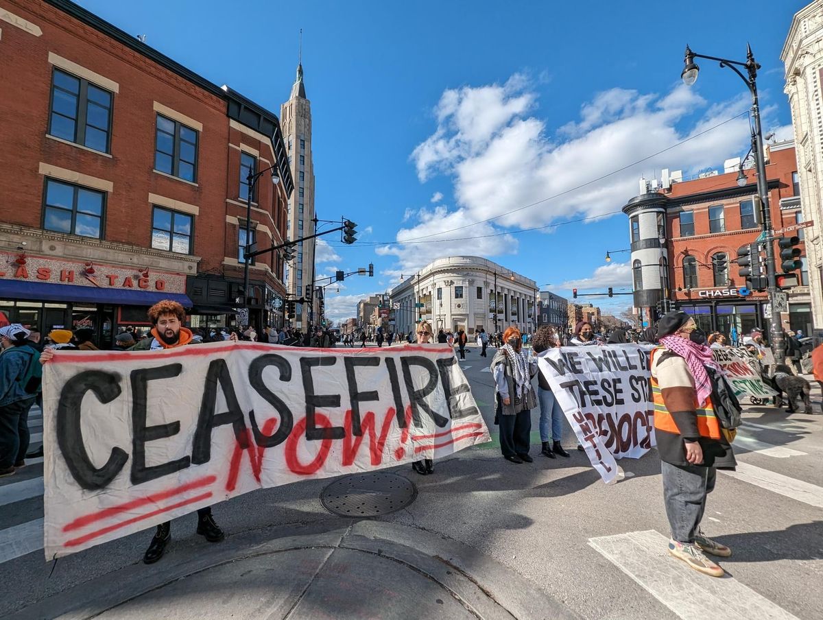 Protesters blockade a three-way intersection holding banners. One reads, "Ceasefire now!"