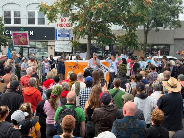 A crowd gathers in front of a banner with a butterfly on it while a man speaks through a mic.