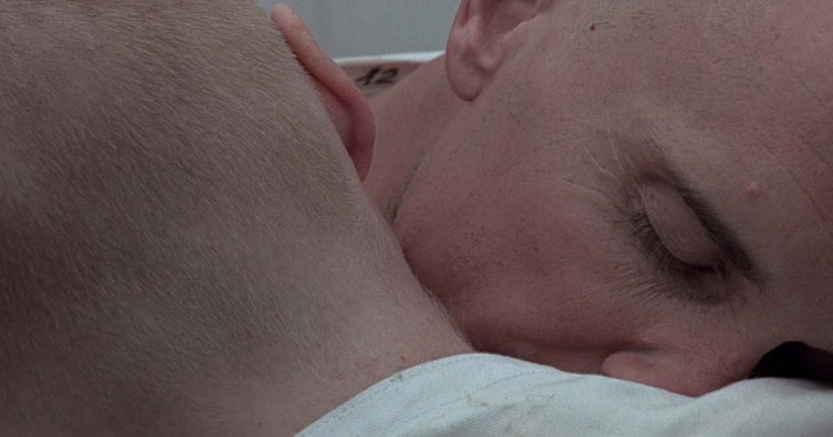 A close-up of two bald characters in white clothing embracing, their heads pressed together in a tender moment.