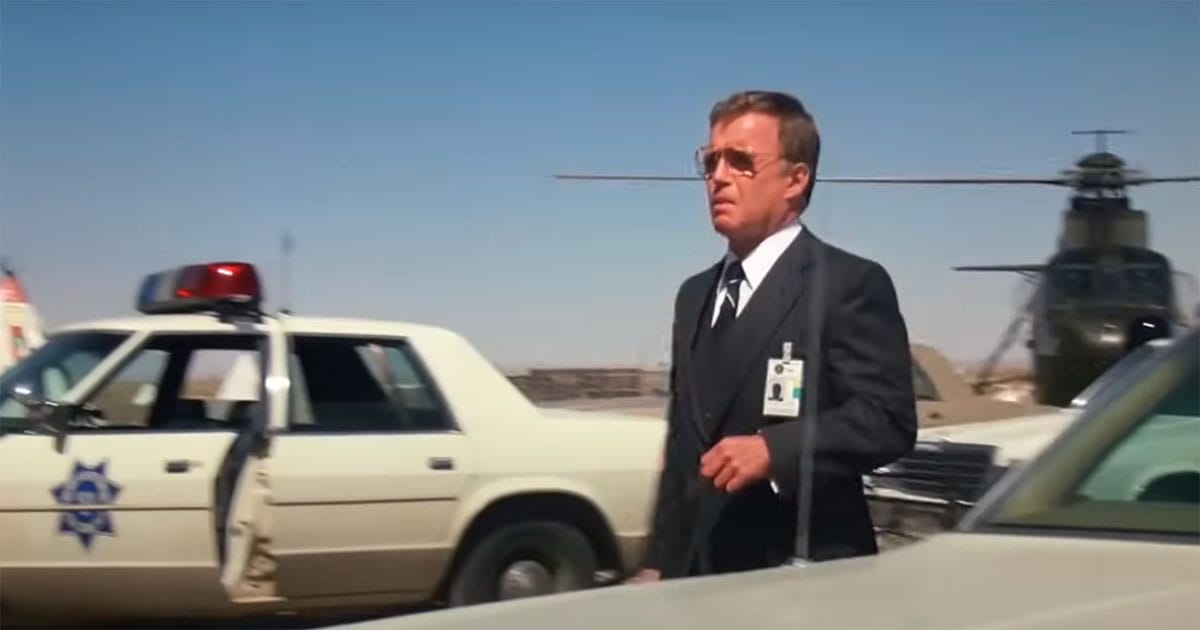 Scene from "Starman" showing a government agent in a suit walking past police cars and a helicopter, reflecting the film’s portrayal of institutional control over alien contact.