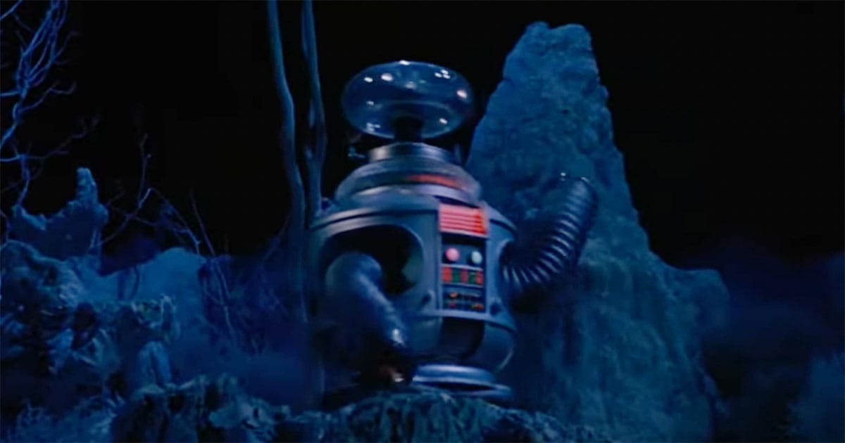 The Robot from "Lost in Space" standing alone in a dark, alien landscape under blue light, evoking mystery and potential danger.