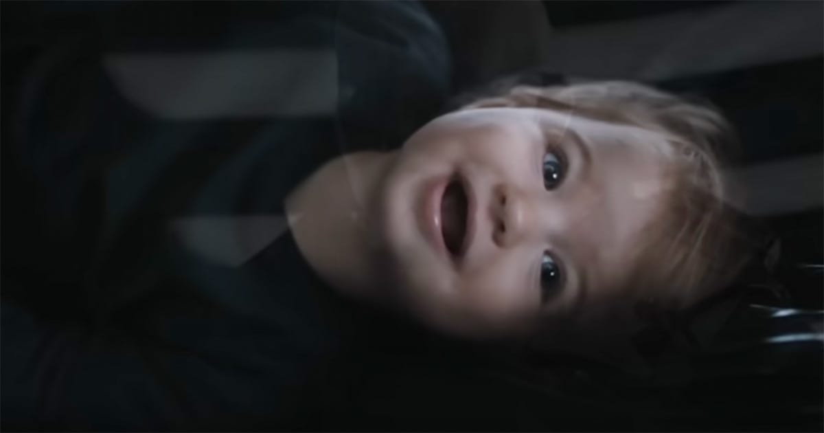 Baby in black clothing smiling under dim lighting with shadowed patterns across her face.