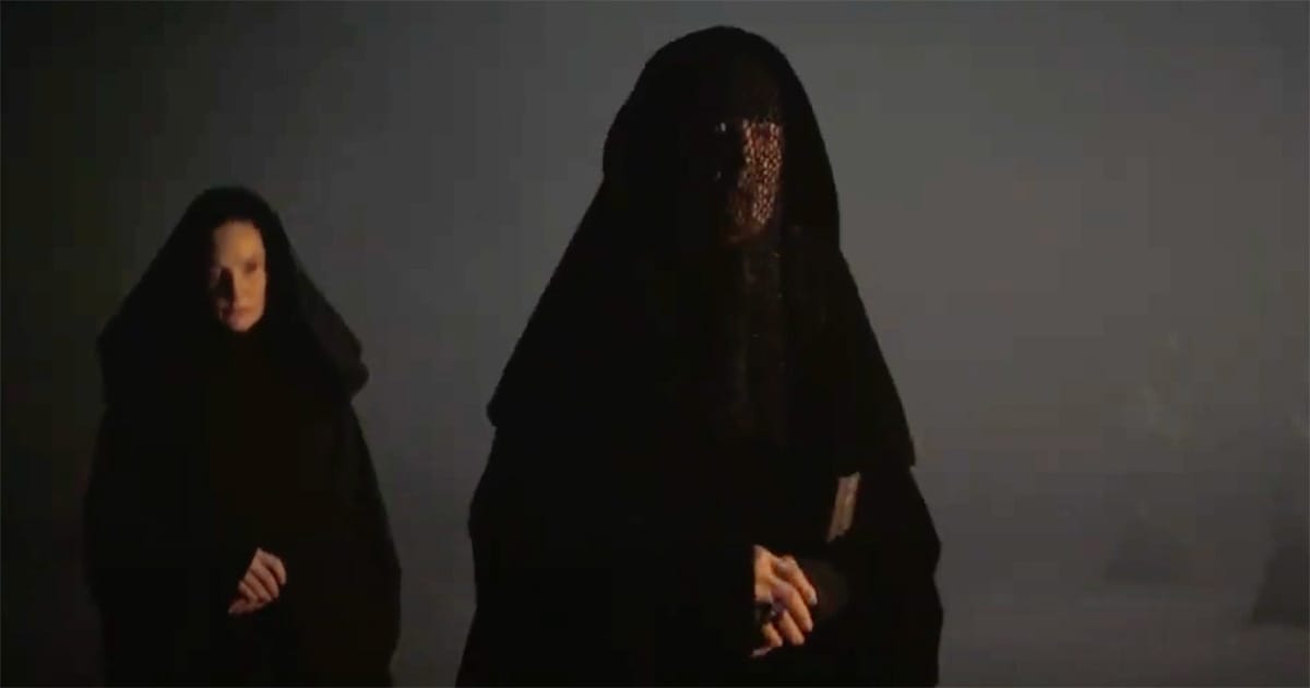 Two Bene Gesserit women in dark robes stand in shadow, one fully veiled and the other dimly lit.