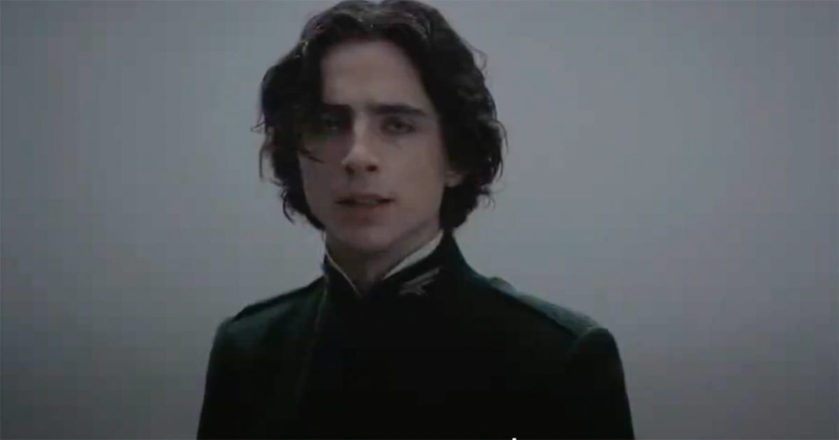 Young man in dark uniform stands in a featureless space, looking ahead with a solemn expression.