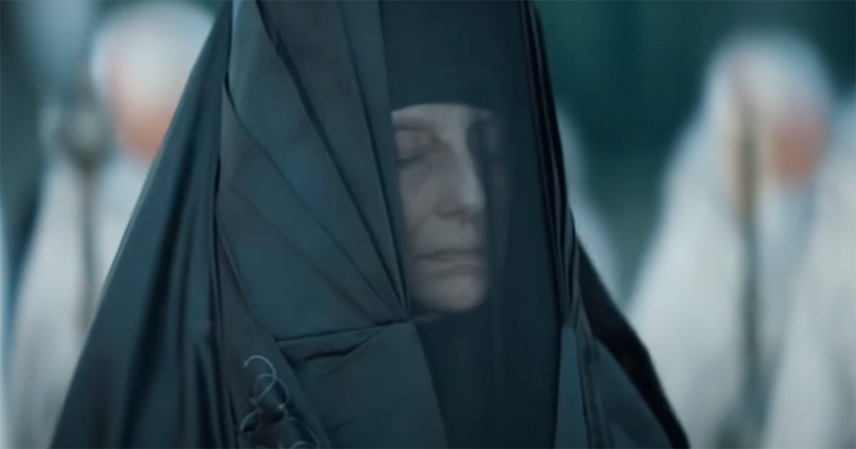 Reverend Mother in dark veil stands with eyes closed among white-robed figures, expression solemn and composed.