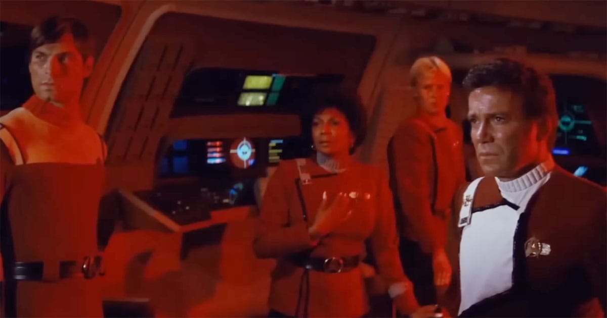 The bridge crew of the Enterprise stands in tense red lighting, with Captain Kirk staring forward, deep in thought, as the crew reacts to unfolding events.