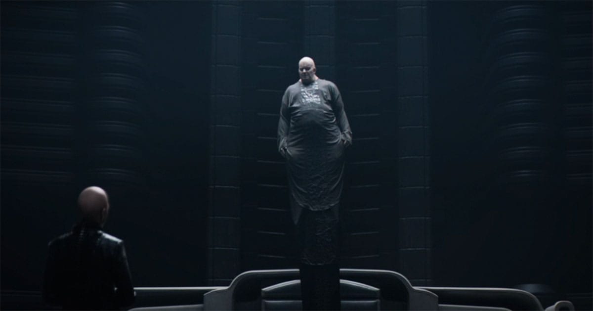 A bald man in long robes hovers midair in a dark hall as another figure stands before him, the scene lit by cold, focused light.