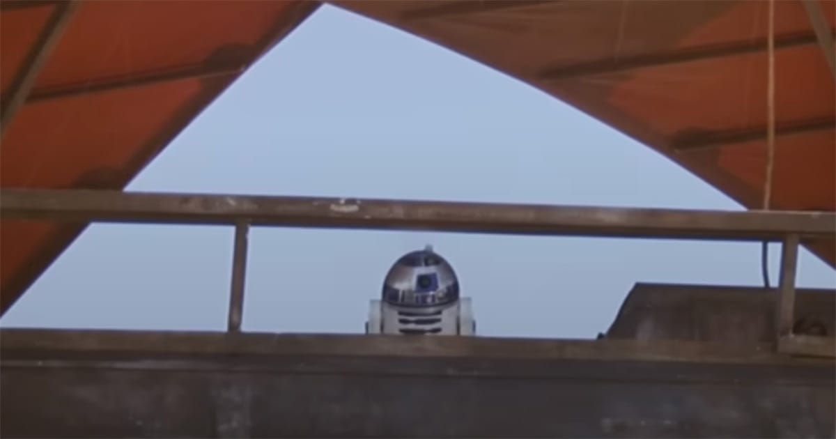 R2-D2 standing on a desert skiff beneath red sails against a bright sky.