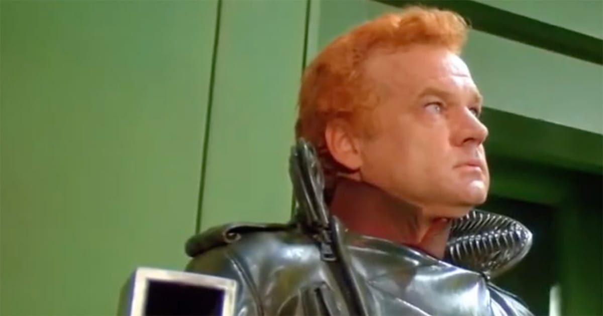 A uniformed officer in black armor stares ahead against a green wall, expression fixed and watchful.