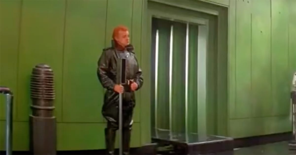 A guard in black uniform stands alone in a wide green corridor, holding a long weapon before an elevator door.