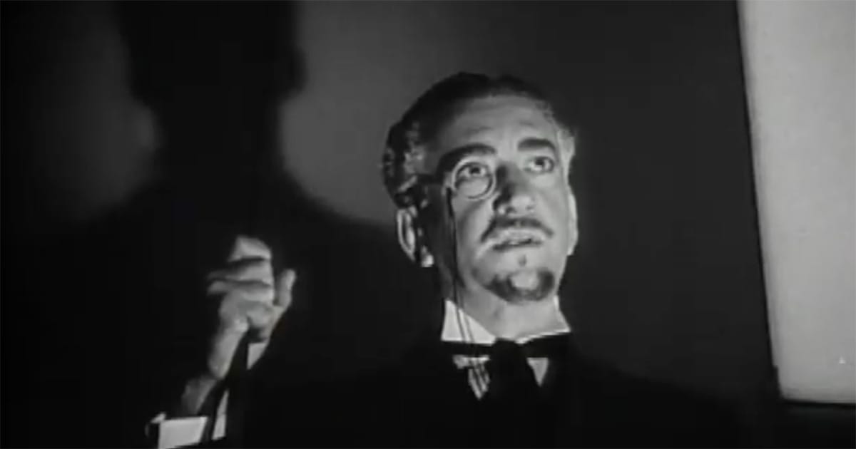 A scientist with a monocle raises his hand as a large shadow falls behind him in a scene from “The Return of Doctor X.”