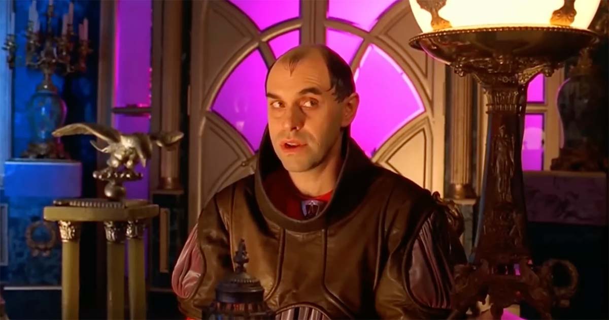 Count Hasimir Fenring seated alone in an imperial interior, facing forward with a composed expression and illuminated architectural details behind him.