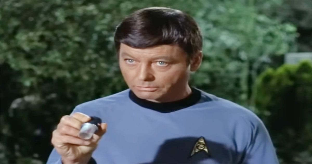 Dr. McCoy holding a small handheld medical device during a Star Trek The Original Series field scene, wearing a blue Starfleet uniform and examining the instrument.