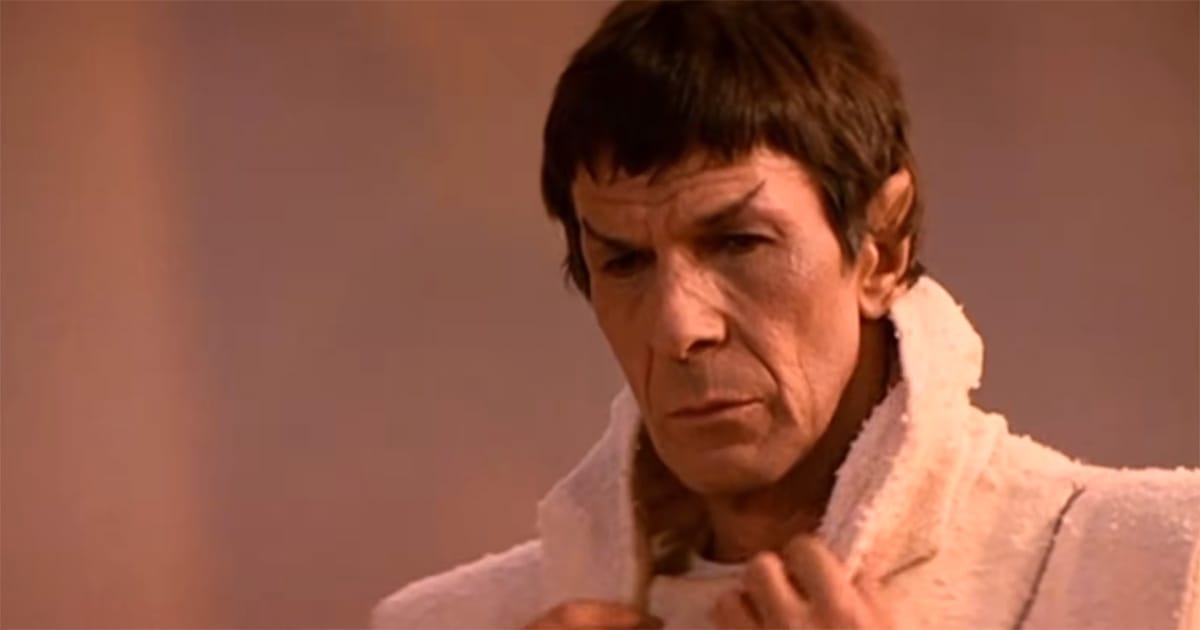 Spock stands in white ceremonial attire with a solemn expression in a formal setting.