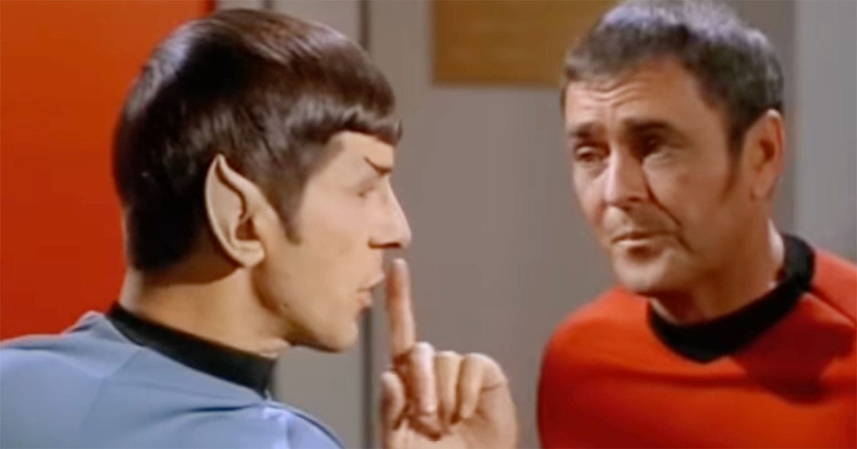 Spock signals for silence with a raised finger while speaking to Montgomery Scott aboard the Enterprise.