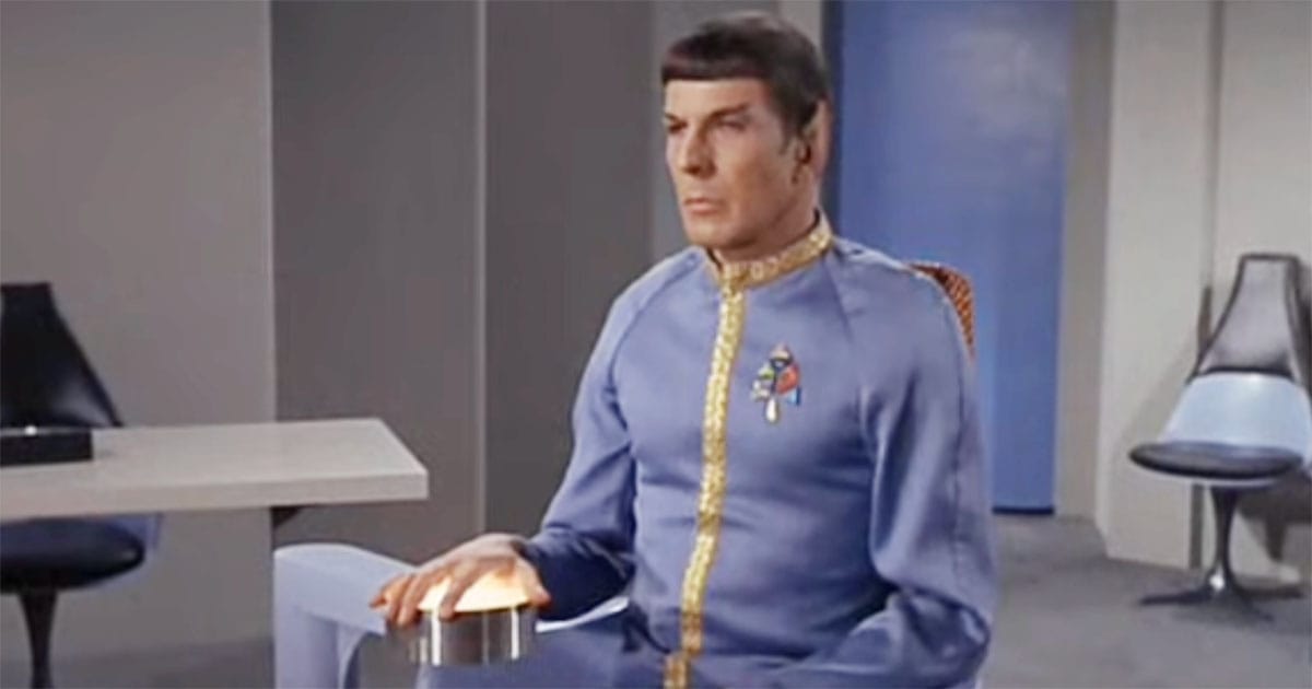 Spock sits composed in a blue uniform during a formal court proceeding with his hand resting on a control device.