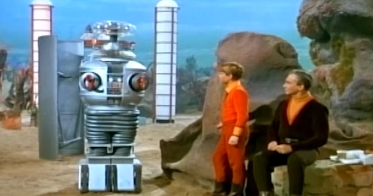 Color scene from "Lost in Space" showing Will Robinson standing beside the B-9 Robot outdoors, with Dr. Smith nearby, illustrating their established companionship.