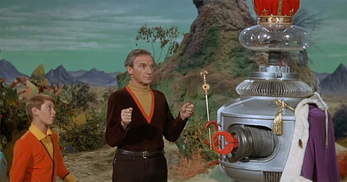 Color scene from "Lost in Space" showing Dr. Smith gesturing toward the B-9 Robot adorned with a crown, with Will Robinson nearby, suggesting the Robot’s symbolic importance.