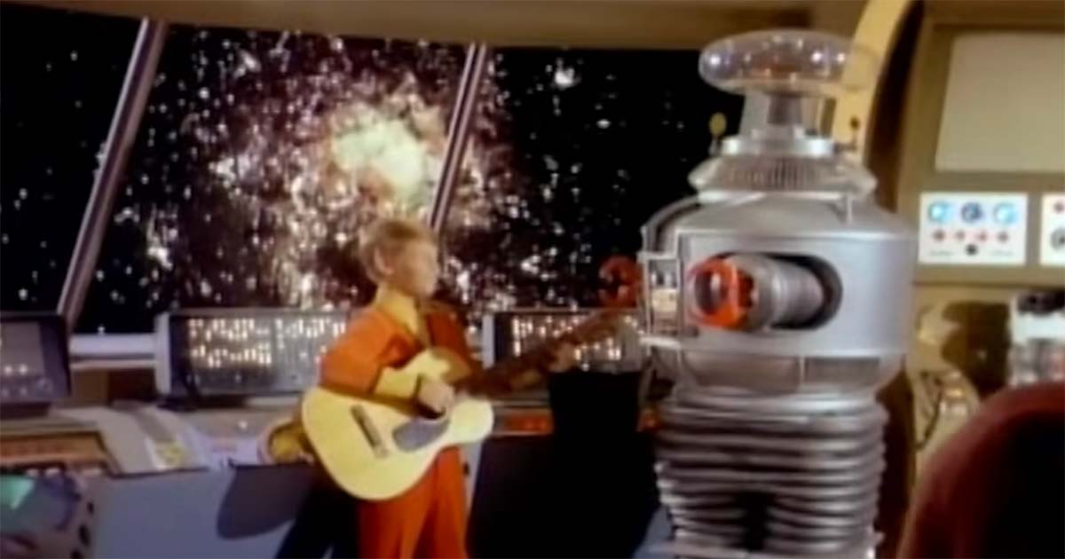 Color scene from "Lost in Space" showing Will Robinson playing guitar to the B-9 Robot inside the spaceship, highlighting their growing relationship and emerging identity.