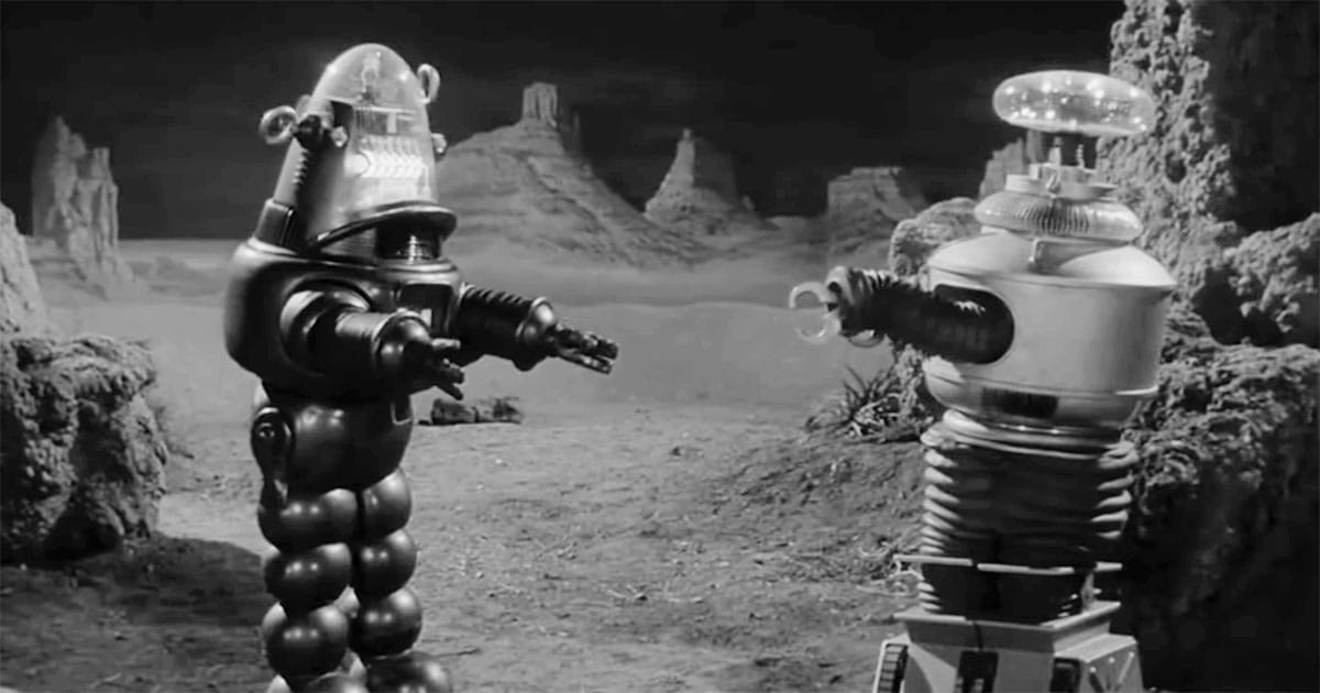 Black-and-white scene from "Lost in Space" showing the B-9 Robot facing another robot on a barren alien landscape, emphasizing their mechanical design and function.