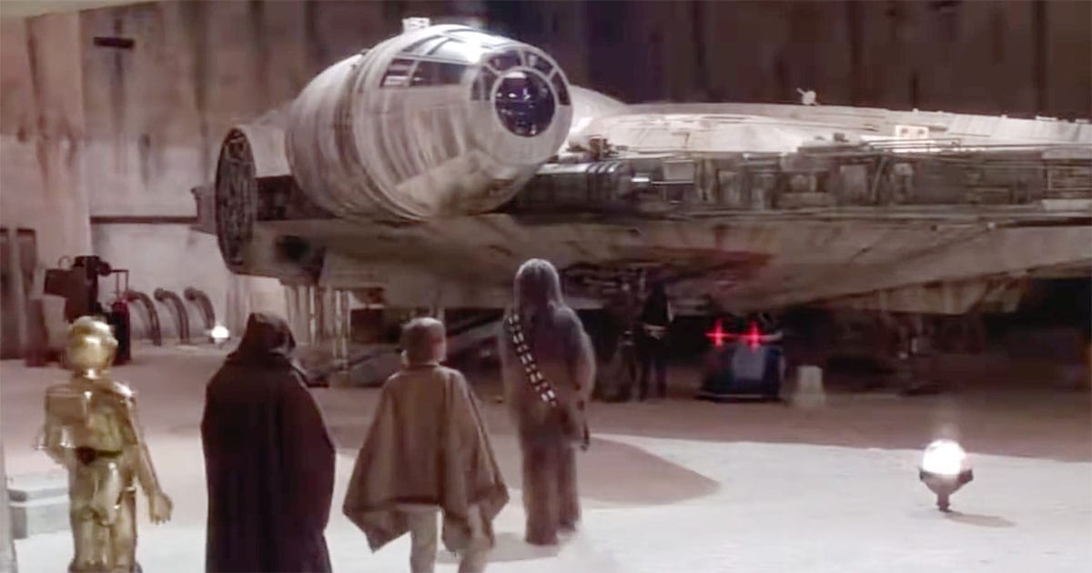 Travelers and two droids stand beside a worn spacecraft in a desert hangar before departing on a journey.