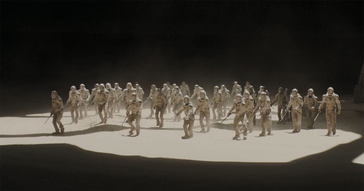 A formation of Sardaukar advances together across a stark illuminated training ground surrounded by darkness.
