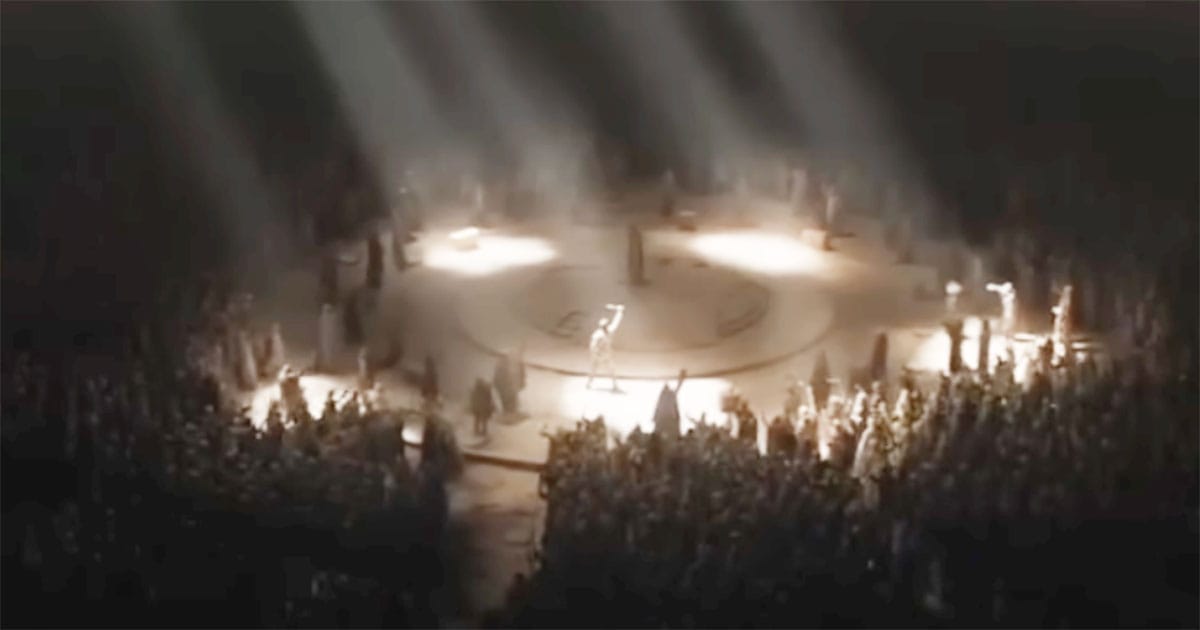 Paul Atreides stands at the center of a large gathering as crowds surround him beneath beams of light.