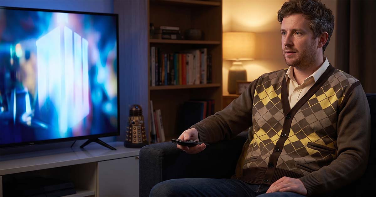 Doctor Who fan wearing a Fourth Doctor-style cardigan while watching the TARDIS on television in a living room with a Dalek collectible nearby.