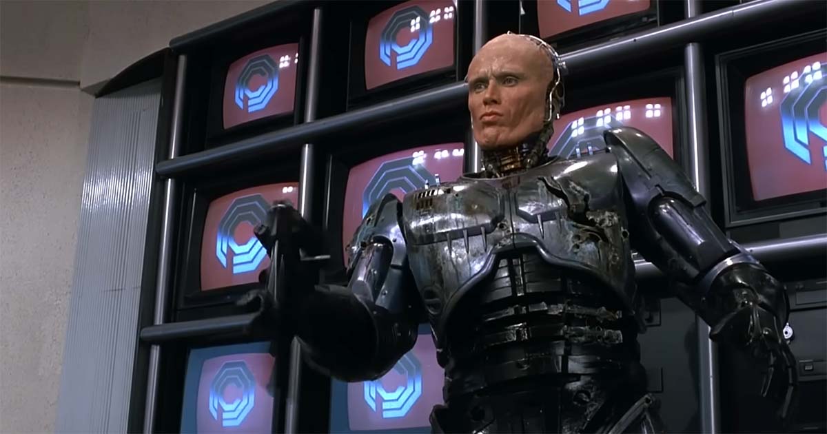 Battle-damaged RoboCop with part of his human face exposed stands before a wall of OCP monitors in a scene from the 1987 science fiction film "RoboCop".