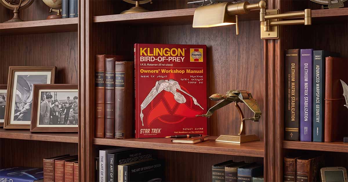 “Klingon Bird-of-Prey Haynes Manual” displayed on a dark wood bookshelf beside a Klingon ship model, books, framed photographs, and a brass library lamp.