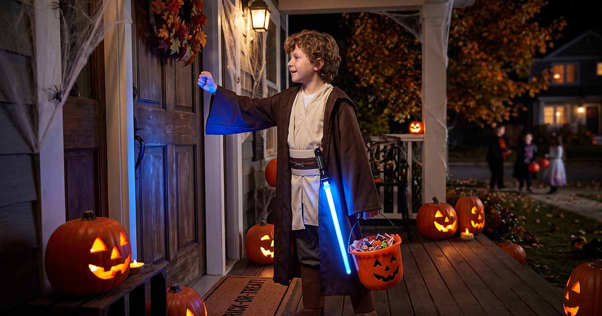 Young Jedi in a brown robe carrying a blue lightsaber and candy bucket while trick-or-treating.