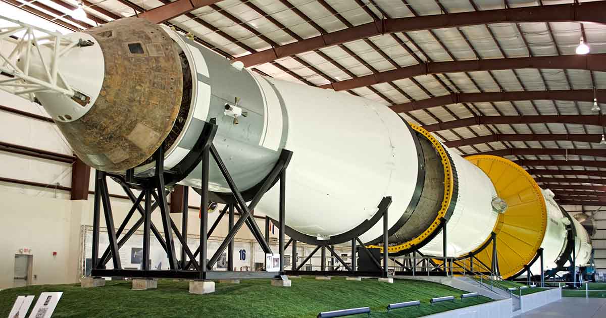 Saturn V Moon rocket displayed horizontally inside a large hangar, showing the enormous scale of the Apollo-era spacecraft.