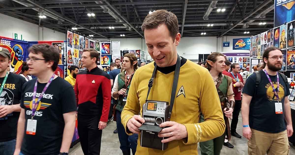 Star Trek fan in a gold command uniform holding a classic tricorder replica at a crowded science fiction convention.