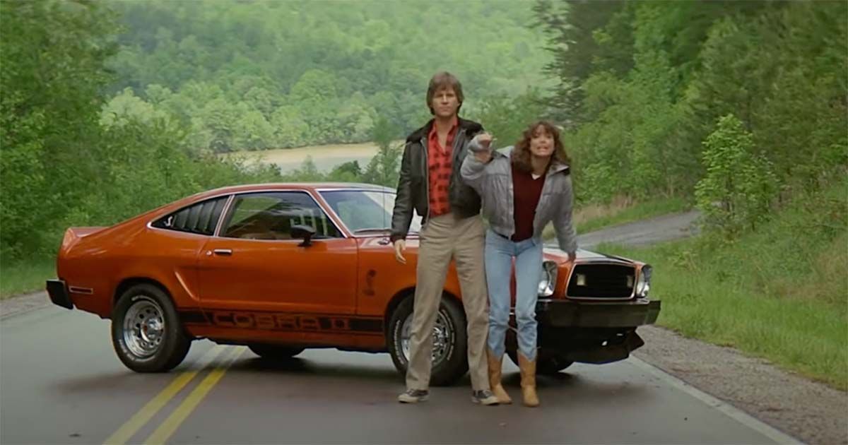 Scene from "Starman" showing Jeff Bridges and Karen Allen standing beside a red Cobra Mustang on a forested road, symbolizing their journey between secrecy and cosmic revelation.