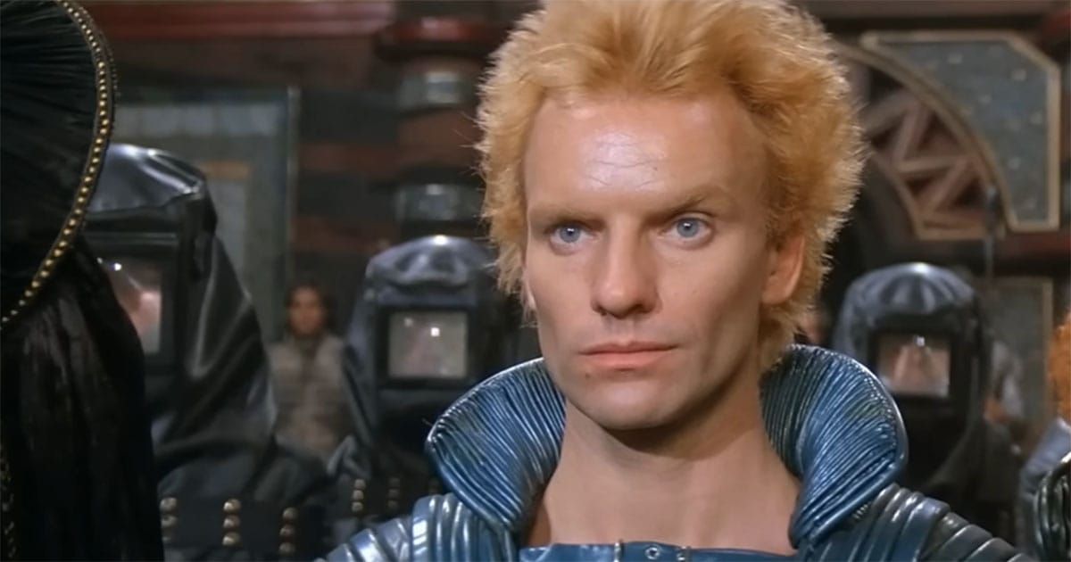Actor Sting as Feyd-Rautha in blue armor, facing forward under harsh lighting with Harkonnen guards behind him.