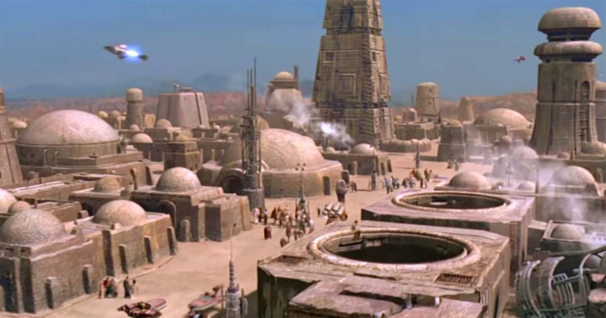 Wide view of Mos Eisley spaceport on Tatooine showing domed buildings, scattered crowds, and ships moving overhead in a worn desert city environment.