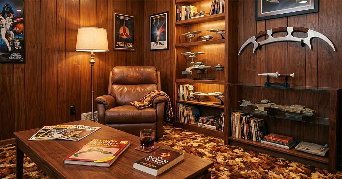 1970s wood-paneled collector’s den with a leather recliner, Klingon books on a coffee table, starship models, a mounted bat’leth, a d’k tahg replica, and warm retro lighting.