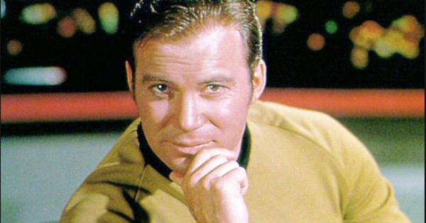 Captain Kirk in a gold Starfleet uniform aboard the USS Enterprise bridge, confidently gazing forward with control panels and ambient lighting in the background.