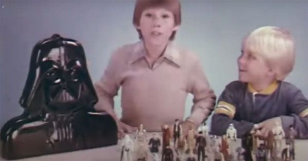 Two children smiling with a Darth Vader-shaped action figure case and Star Wars figures displayed on a table.