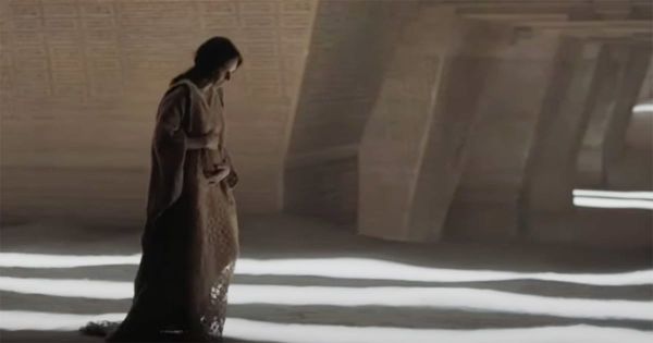 Jessica pregnant, walking through a dimly lit chamber with patterned walls and glowing lines on the sand floor.