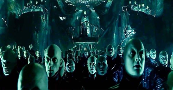 A group of pale, dark-coated beings assemble under green light in an underground chamber from “Dark City.”