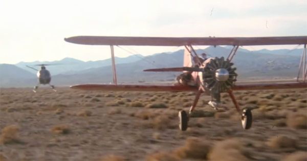 A vintage biplane flying low over a desert while a military helicopter pursues it in a climactic chase scene from the 1977 film Capricorn One.
