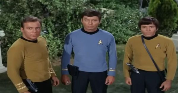 Three Star Trek original series officers standing outdoors with visible phasers and handheld equipment, wearing gold and blue uniforms during a field mission.
