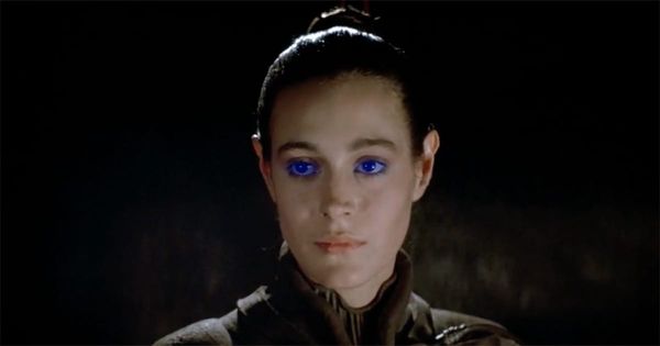 Close-up of Chani from "Dune" 1984 with blue eyes and a calm expression, wearing a stillsuit against a dark background.