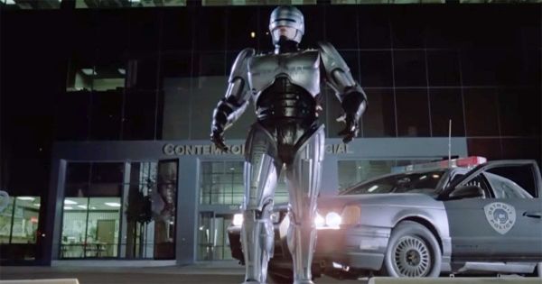 RoboCop stands outside a Detroit police station in his silver cybernetic armor beside a police cruiser in a scene from the 1987 science fiction film "RoboCop".