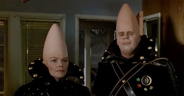 Beldar and Prymaat Conehead stand inside a suburban home wearing formal Remulak uniforms in a scene from "Coneheads" (1993).