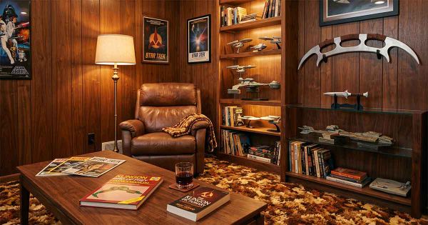 1970s wood-paneled collector’s den with a leather recliner, Klingon books on a coffee table, starship models, a mounted bat’leth, a d’k tahg replica, and warm retro lighting.
