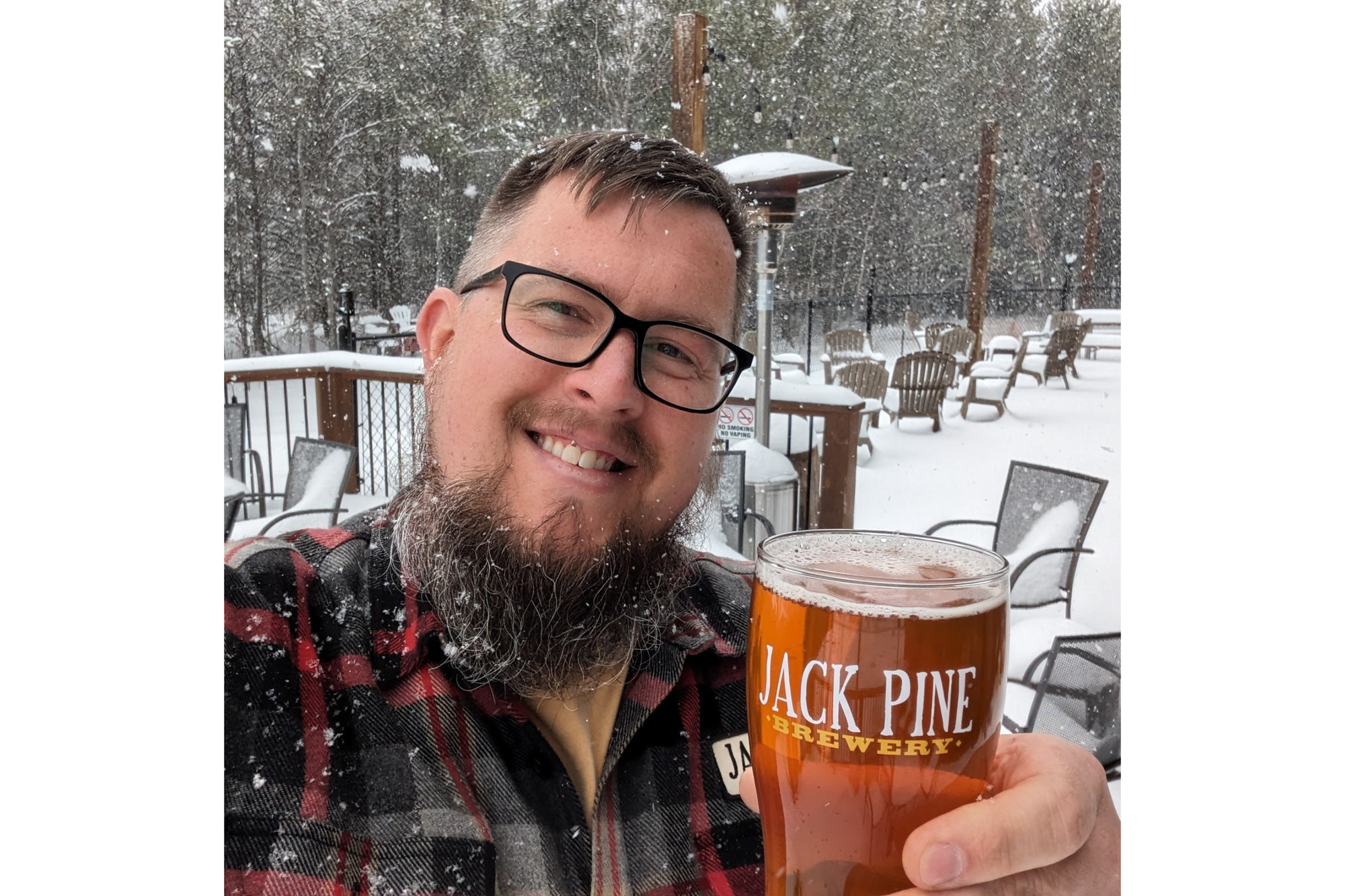 Man with a beard and glasses smiles for a photo. He's holding a glass of beer in a snowy backdrop.
